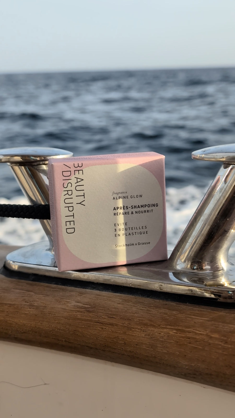 A pink and white box of Beauty Disrupted Apres-Shampooing hair care product resting on a wooden surface next to shiny metal boat cleats, with the ocean in the background.