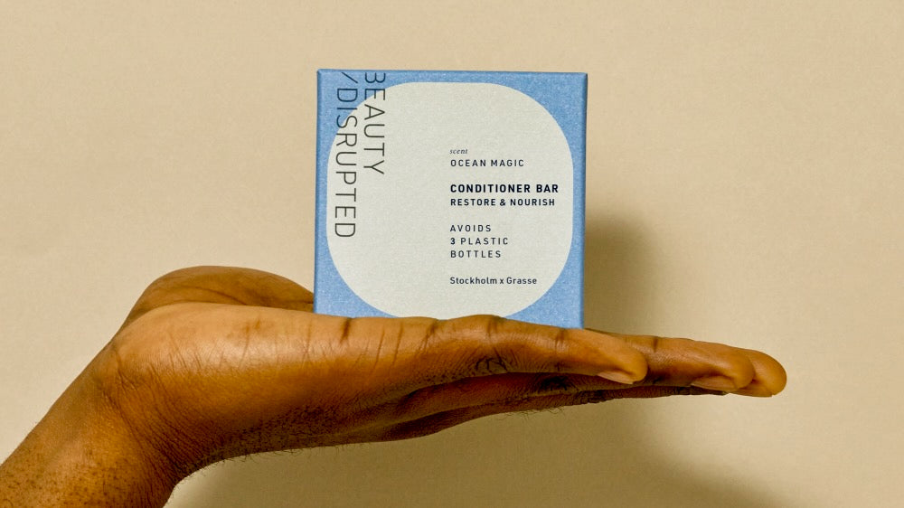 Beauty Disrupted Ocean Magic solid conditioner bar packaging displayed on an open hand against a warm neutral background. Boîte de l’après-shampooing solide Ocean Magic de Beauty Disrupted présentée sur une main ouverte, sur un fond neutre et chaleureux.