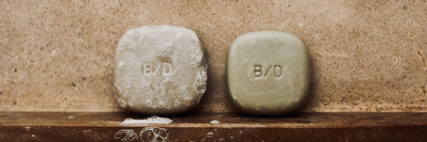 Beauty Disrupted solid shampoo bars on a wet shower ledge, showing lather and the B/D logo embossed on the bars.