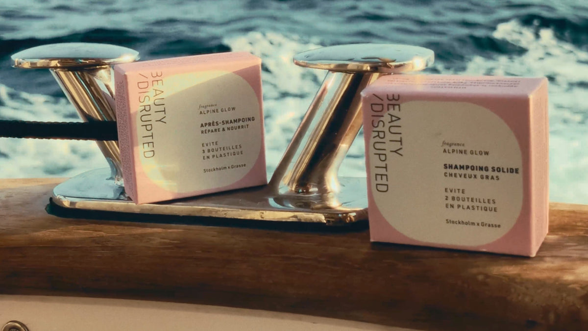 Two pink boxes of Beauty Disrupted Alpine Glow shampoo and conditioner placed on a wooden surface between two shiny metal mooring posts, with the ocean water in the background.